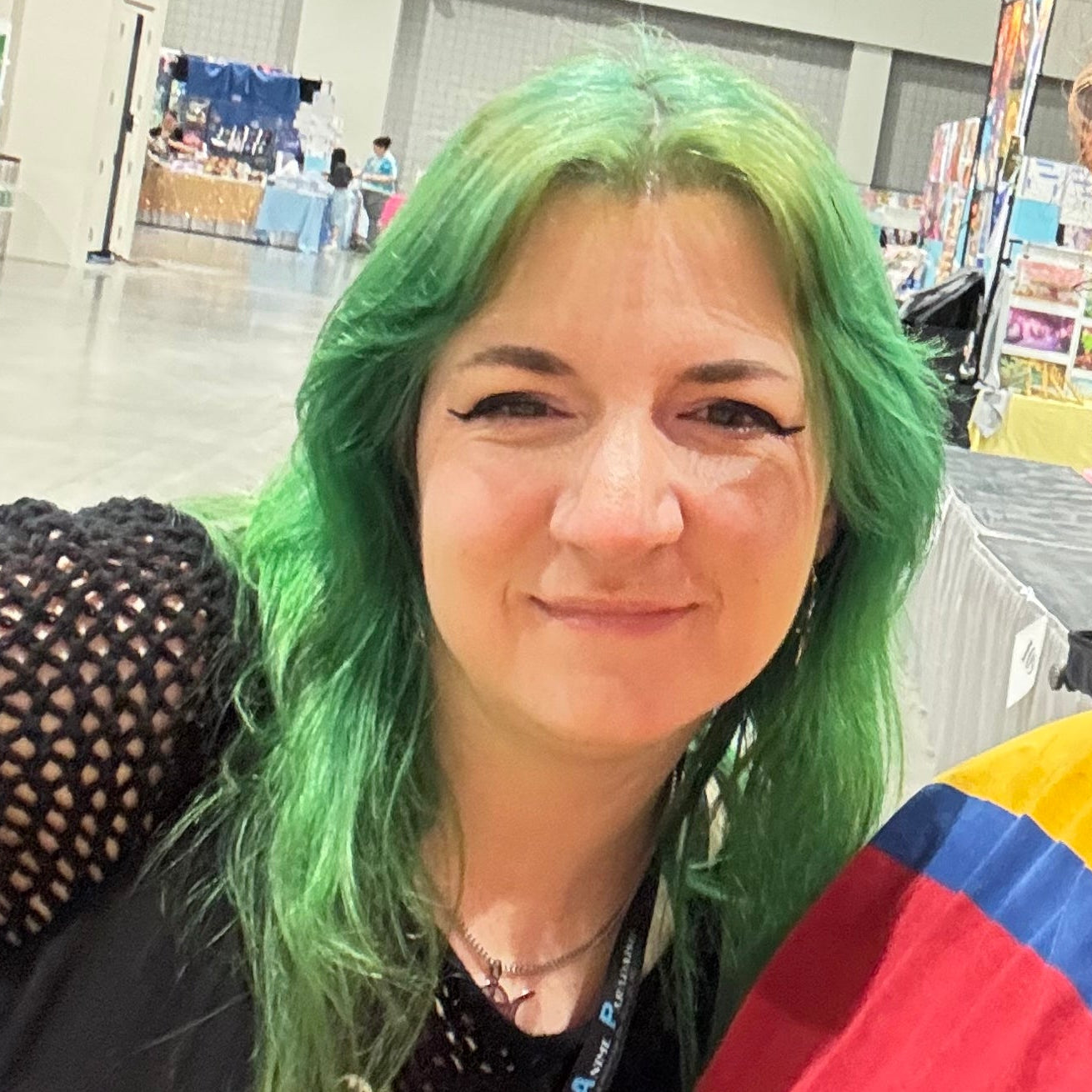 Emi the convention booth helper with green long hair and a black outfit taking a selfie and smiling at the camera