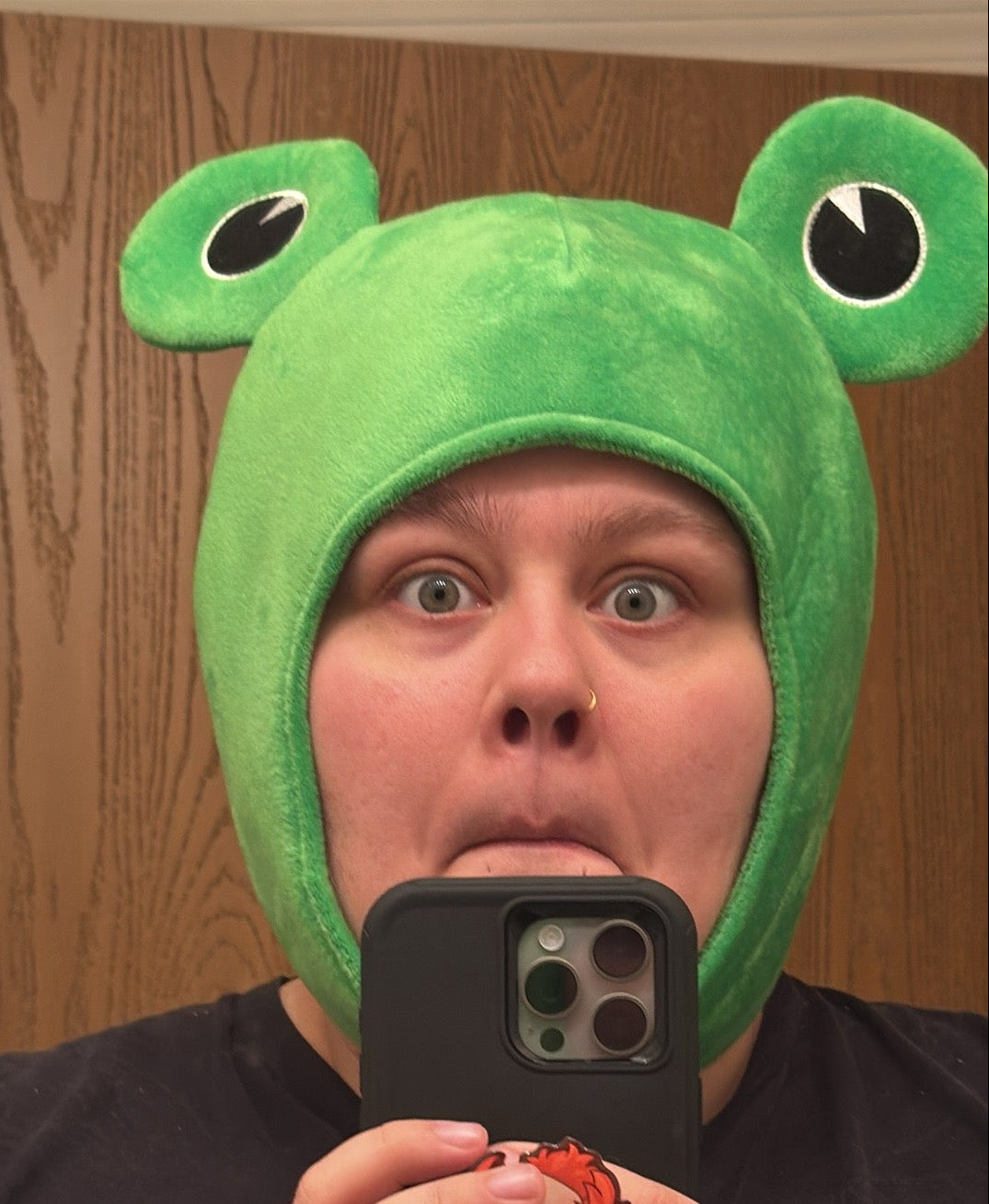 Howl, a booth runner for Mintimeli, taking a mirror selfie wearing a bright green plush frog hood hat with large cartoon eyes and holding a black smartphone in front of their face.